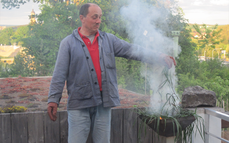 Man sieht Dieter Wolfahrt, wie er das Gesicht verziehend, unerwünschte Beikräuter in ein Feuer im Grill legt.